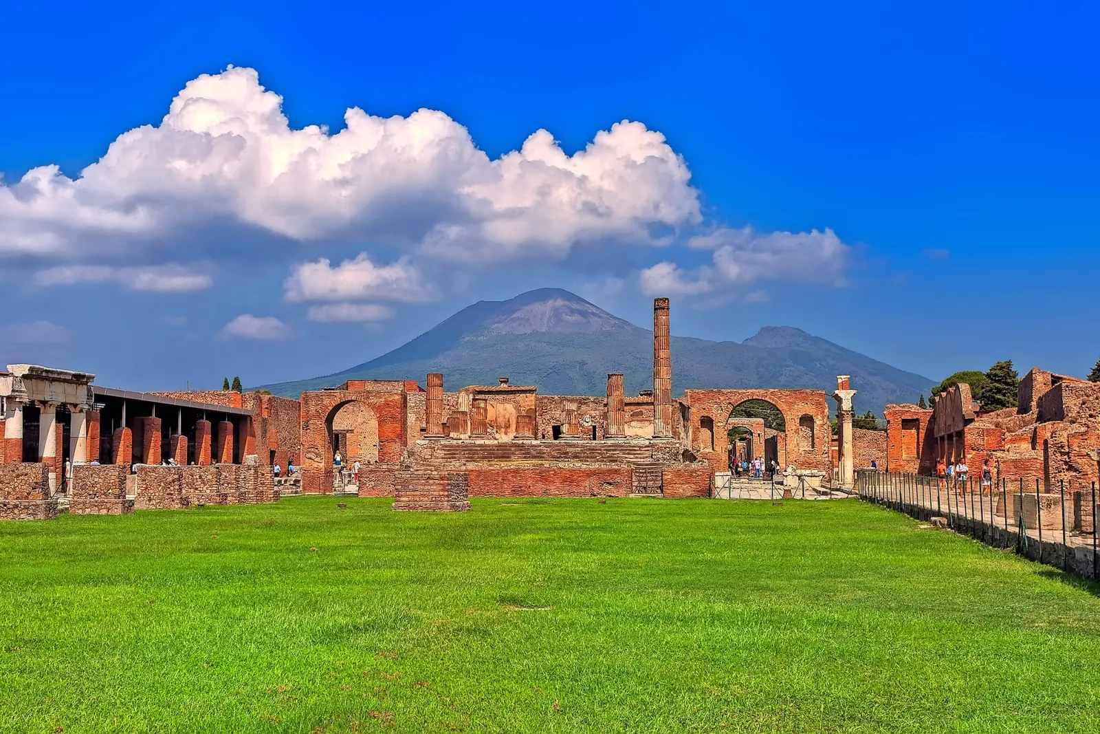 Ruins of Pompeii in front of Vesuvius with green grass in the foreground and blue sky.