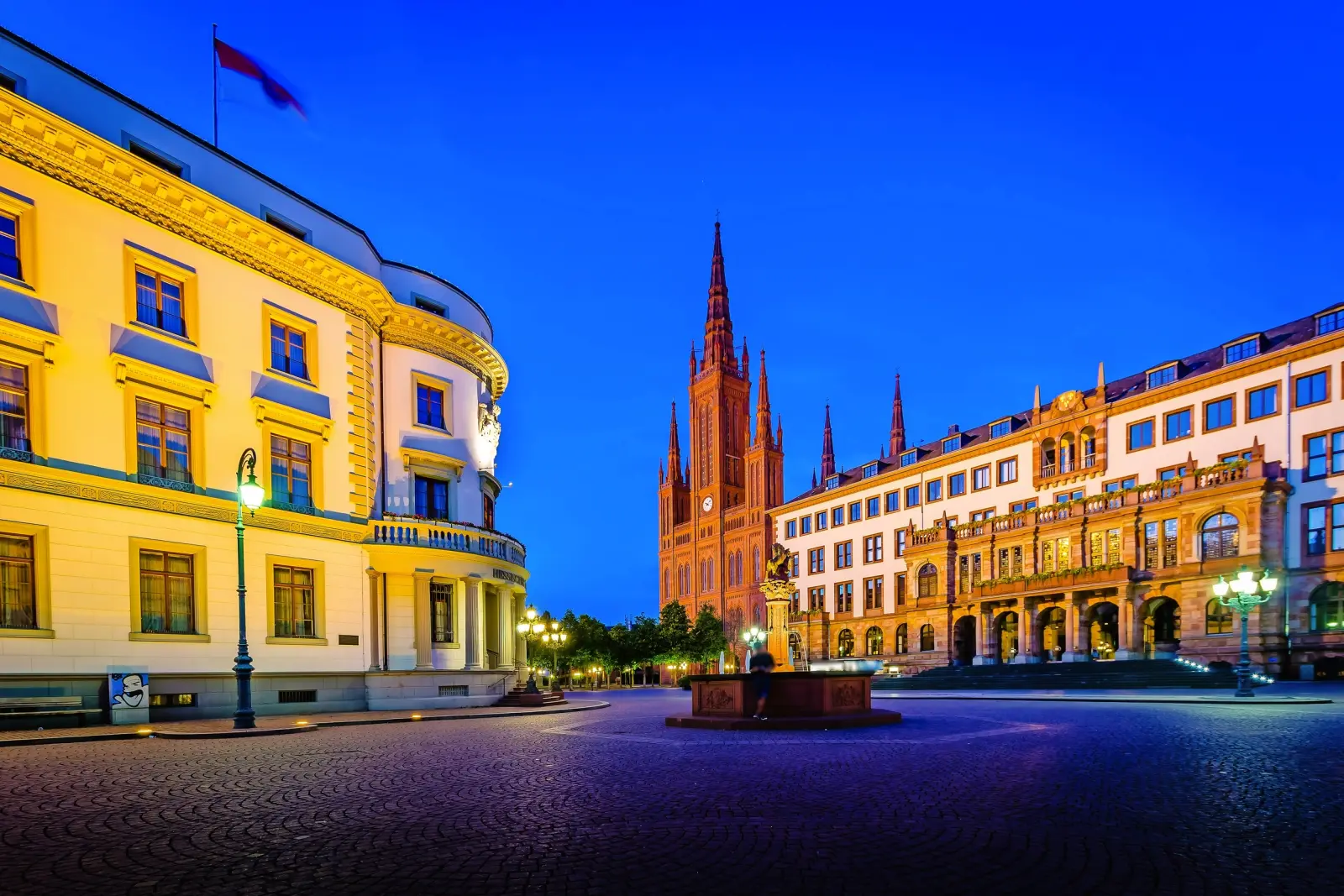 Piazza cittadina di notte con architettura storica, a sinistra un edificio illuminato con bandiera, sullo sfondo una chiesa gotica dalle alte torri, circondata da altri edifici storici.