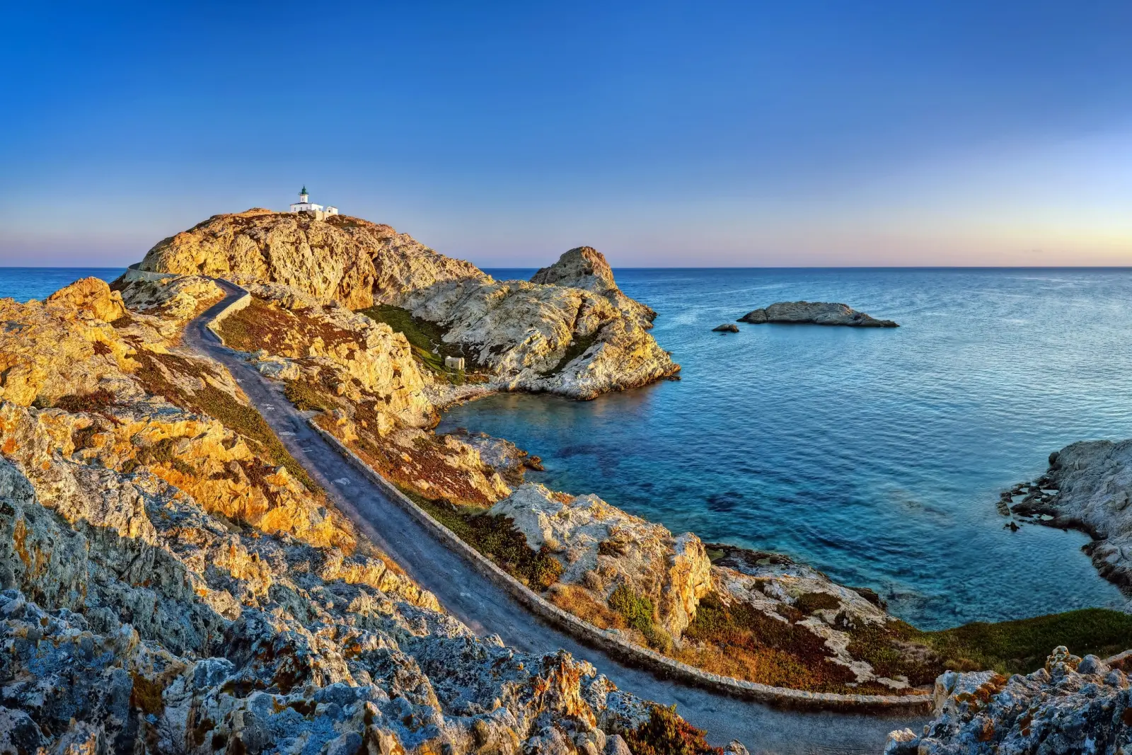 Coastal landscape at sunset with a lighthouse on rocks above the sea.