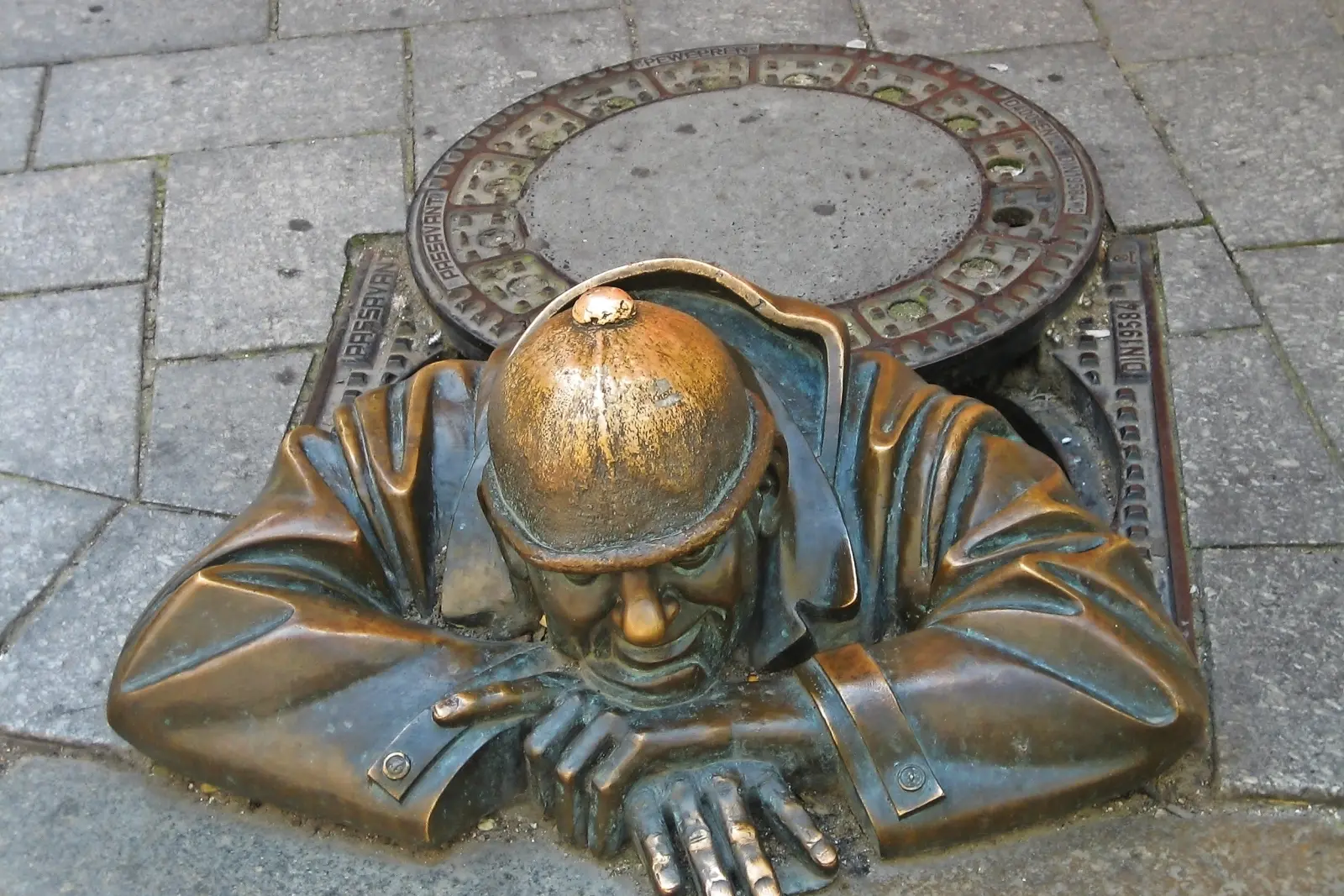 Bronzestatue eines Mannes mit Helm, der aus einem Gully-Deckel auf dem Gehweg schaut.
