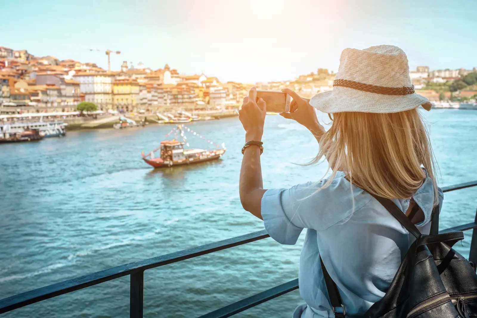 A woman photographing a boat on a river with a city in the background.