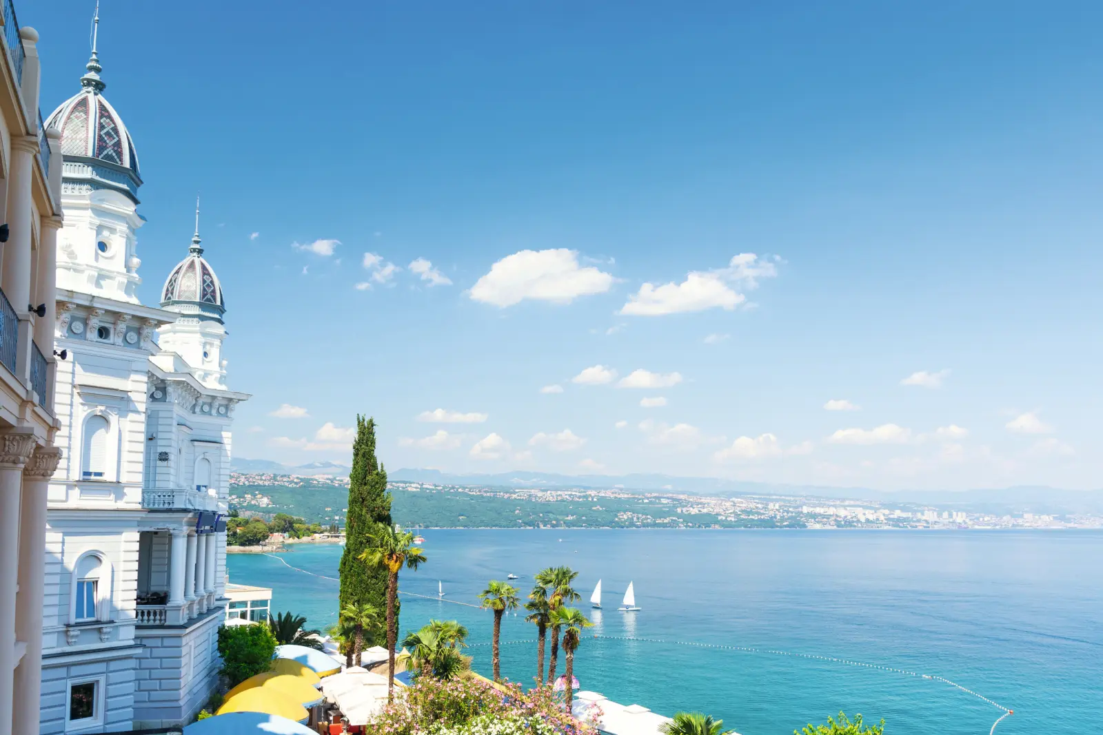 Paesaggio costiero con edificio storico, palme e barche a vela su mare blu.