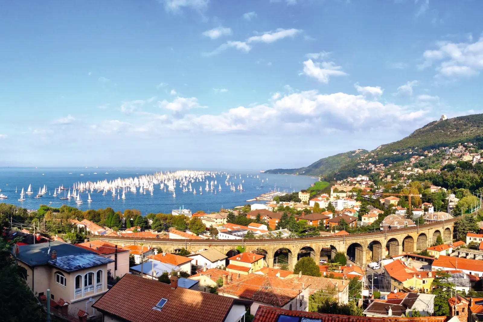 Città sul mare con molte barche a vela e colline sullo sfondo.