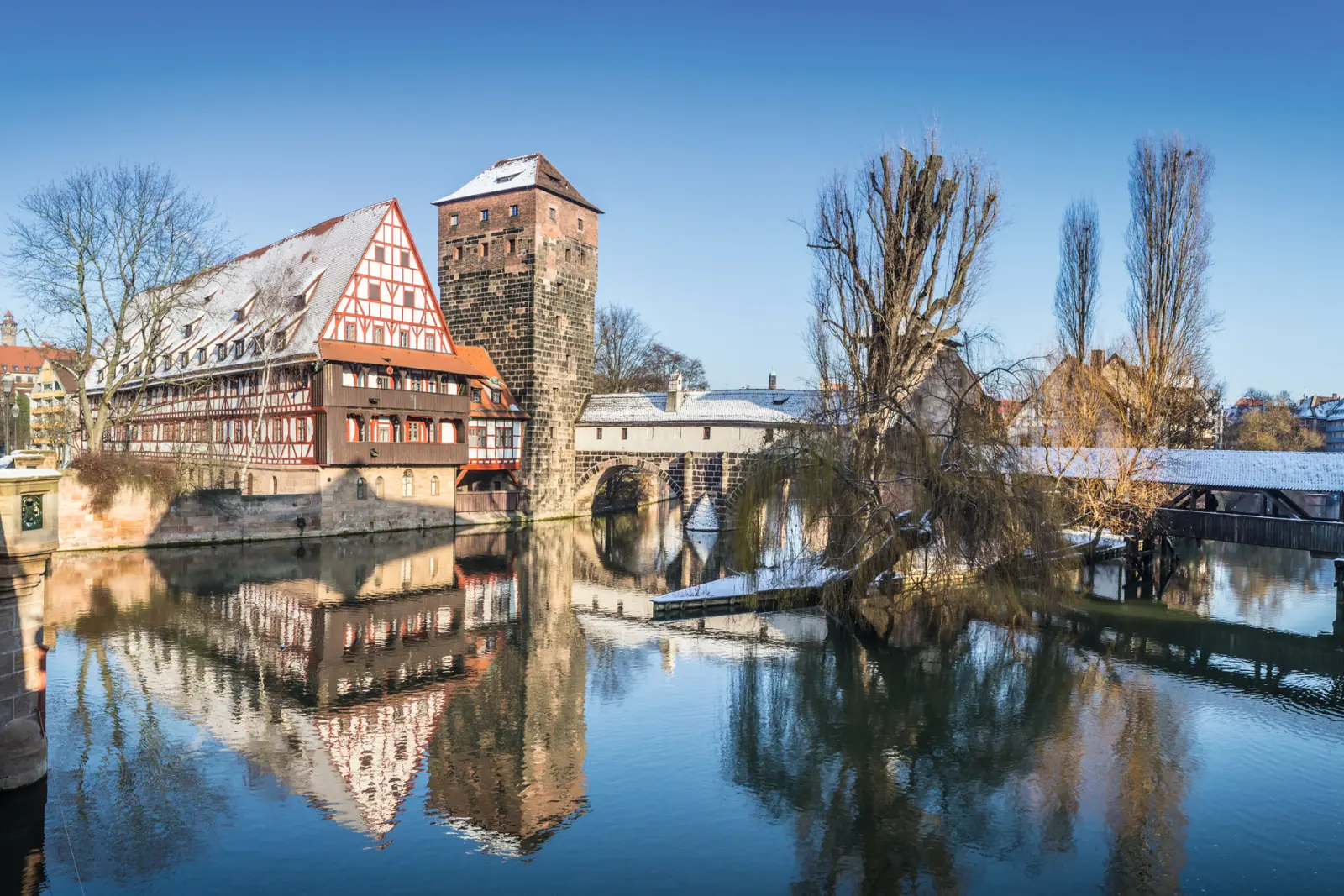 Half-timbered house and bridge on a snowy riverbank, clear sky, reflective water.