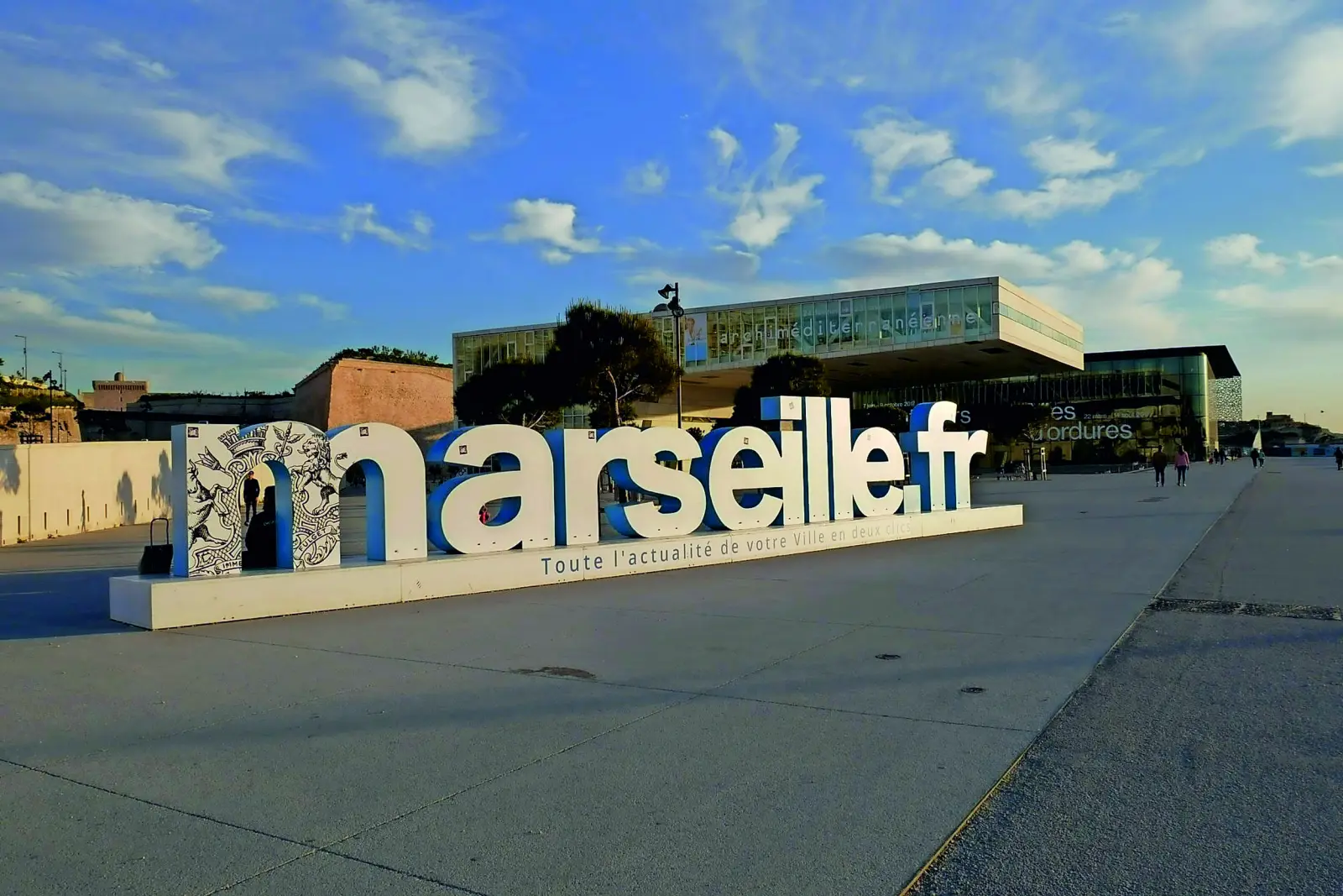 Große Schriftzüge "marseille.fr" vor modernem Gebäudekomplex bei Sonnenschein.