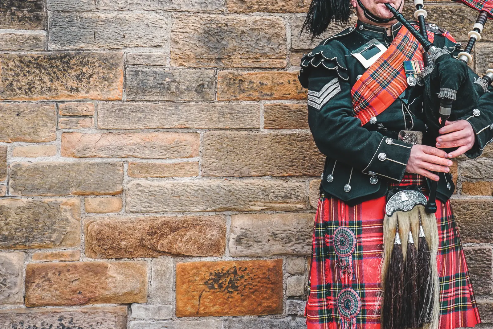 Joueur de cornemuse en kilt devant un mur de pierre.