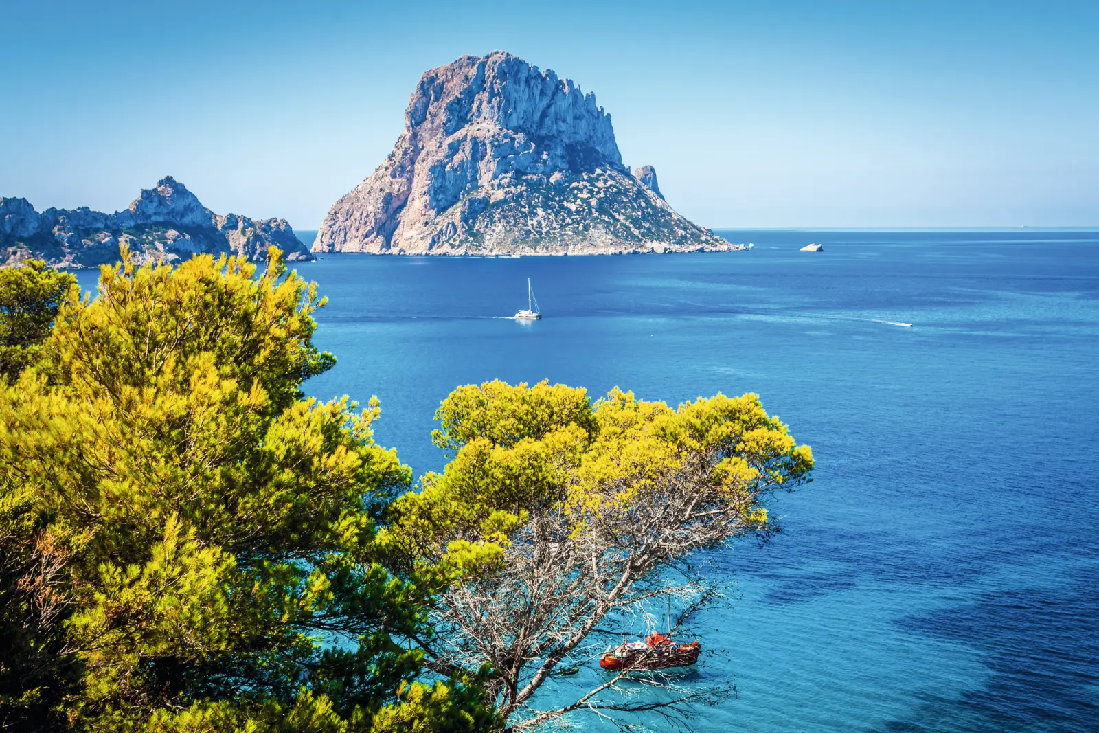 Blick von Cala d'Hort auf die Felseninsel Es Vedrà bei strahlend blauem Himmel und klarem Wasser.