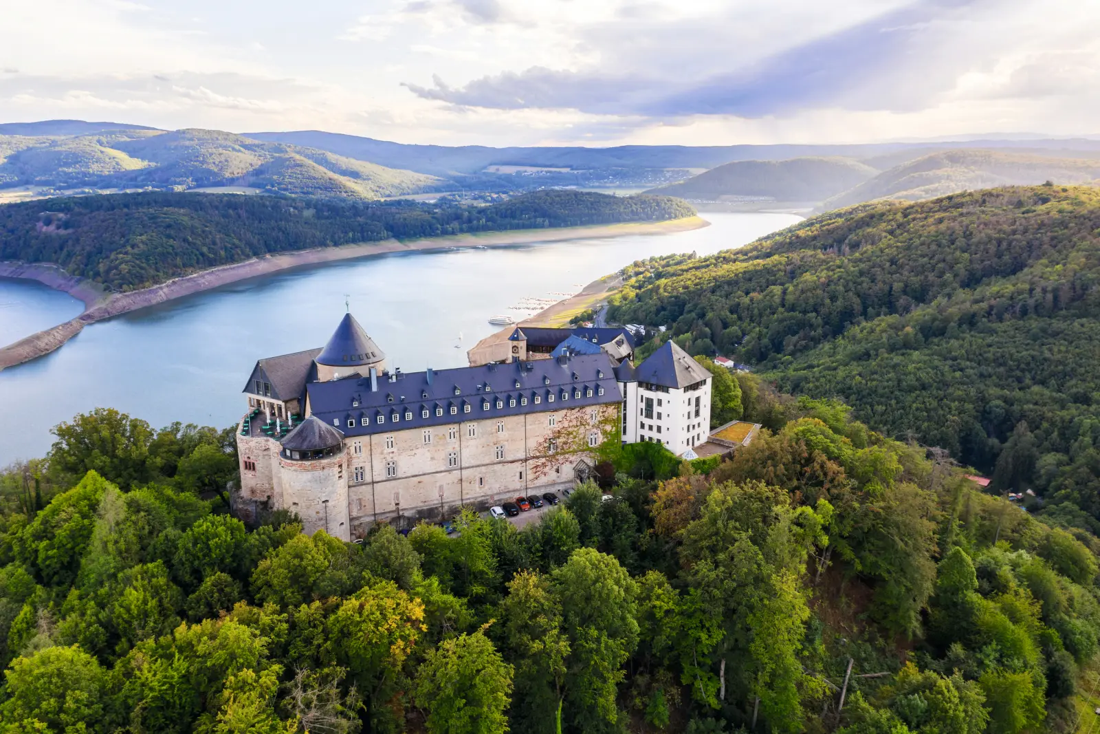 Vue aérienne d'un château historique entouré de forêts et d'un lac, dans un paysage vallonné