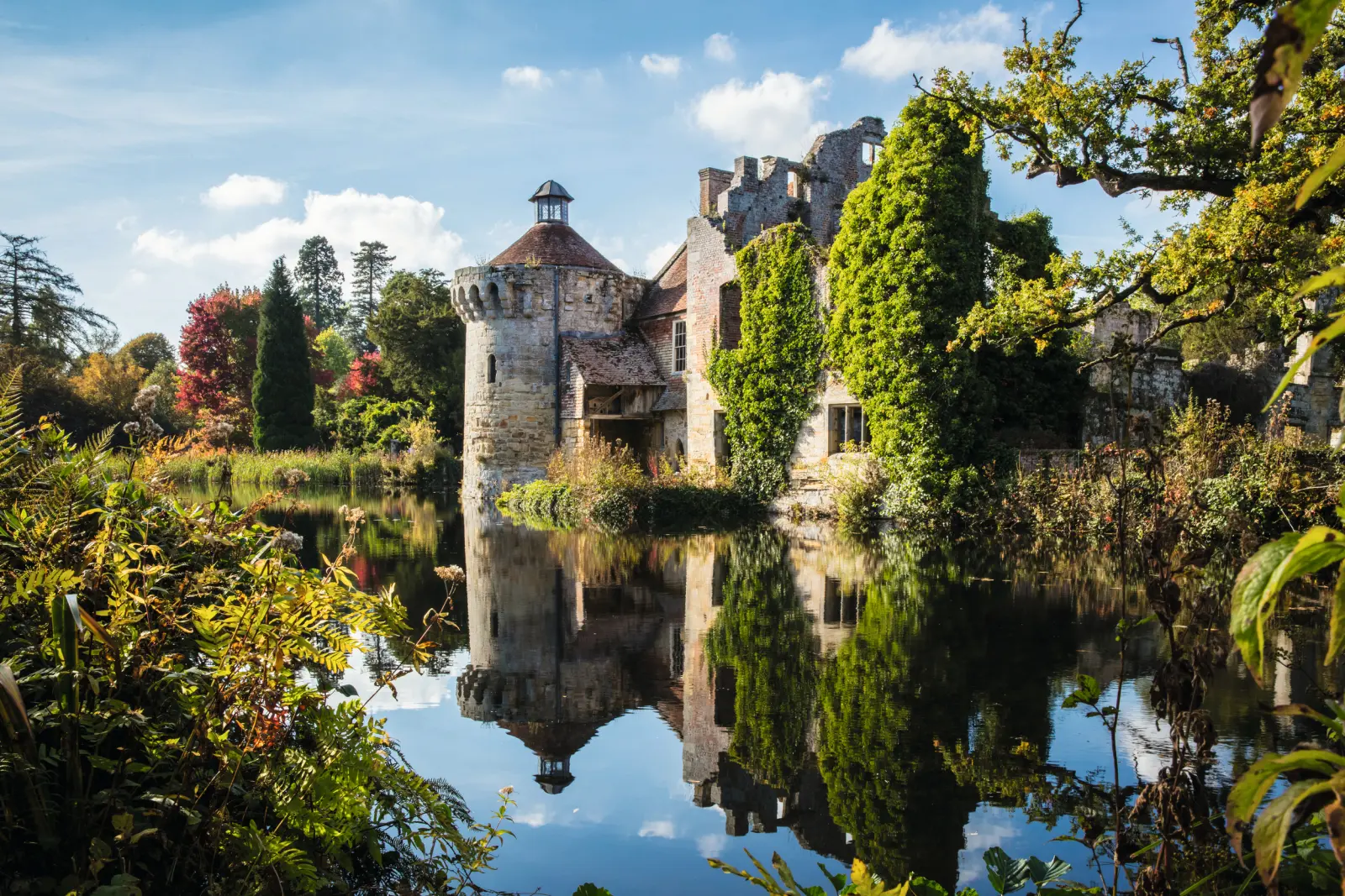 Medieval castle with greenery on the riverbank, autumnal trees in the background.