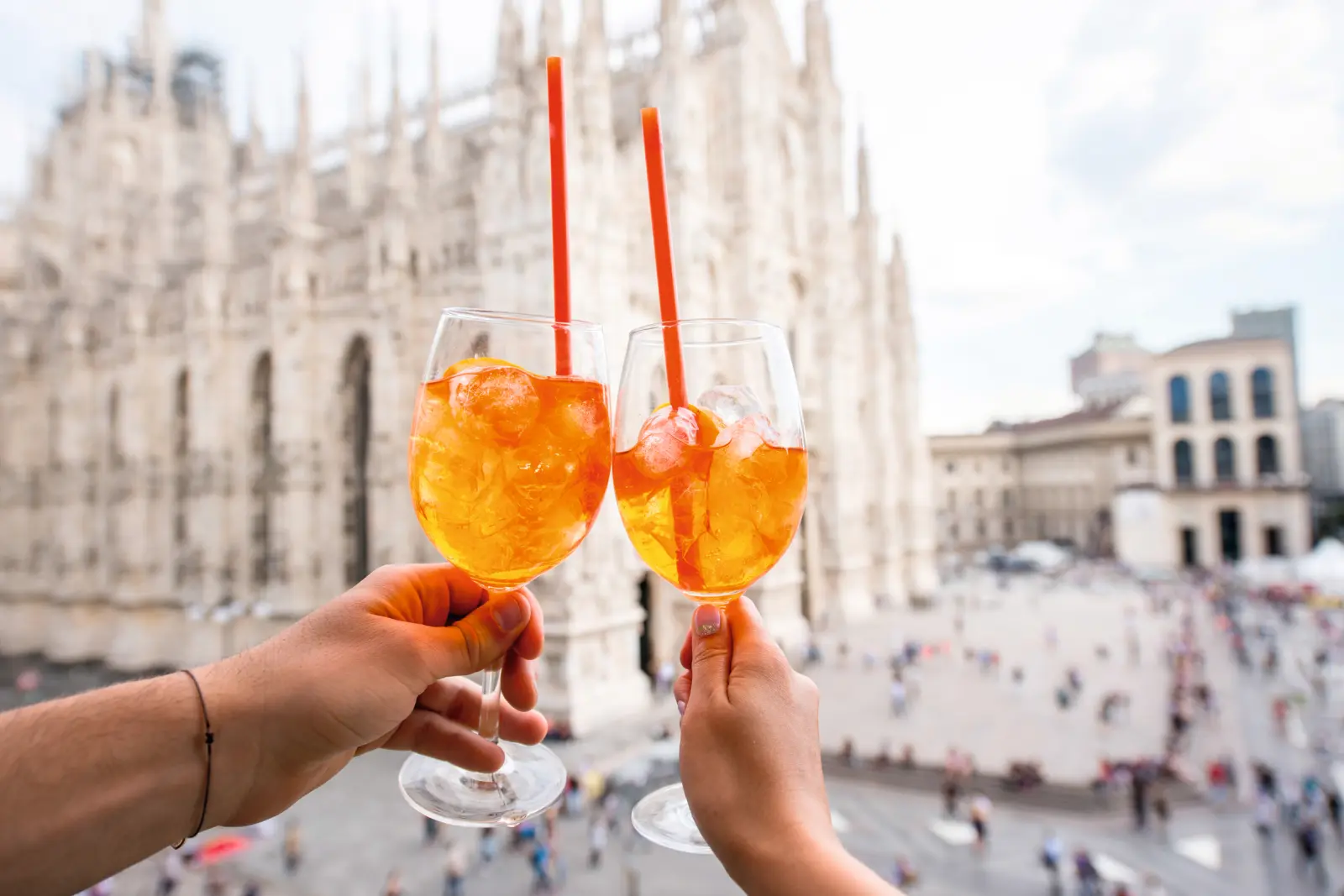 Glasses of Aperol Spritz being raised in a toast in front of Milan Cathedral on the Piazza del Duomo.