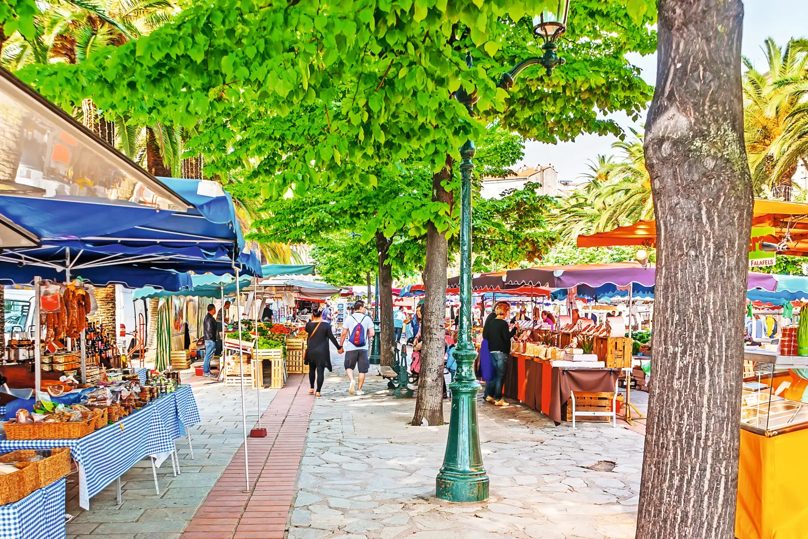 Bancarella del mercato sotto gli alberi, con visitatori e ombrelloni colorati.