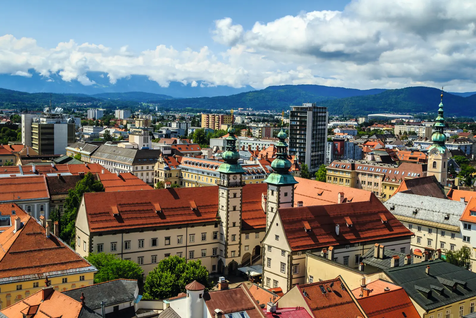 Stadtansicht mit historischen Gebäuden, roten Dächern und Bergen im Hintergrund.