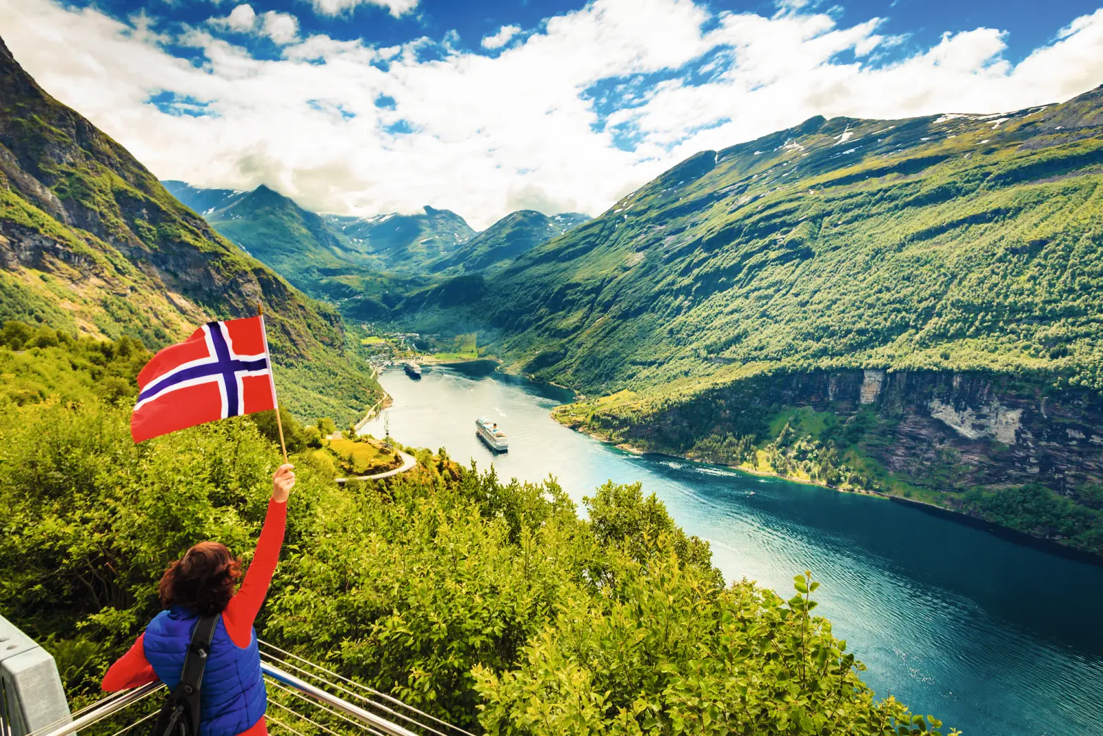 Person schwenkt norwegische Flagge vor Fjordlandschaft.