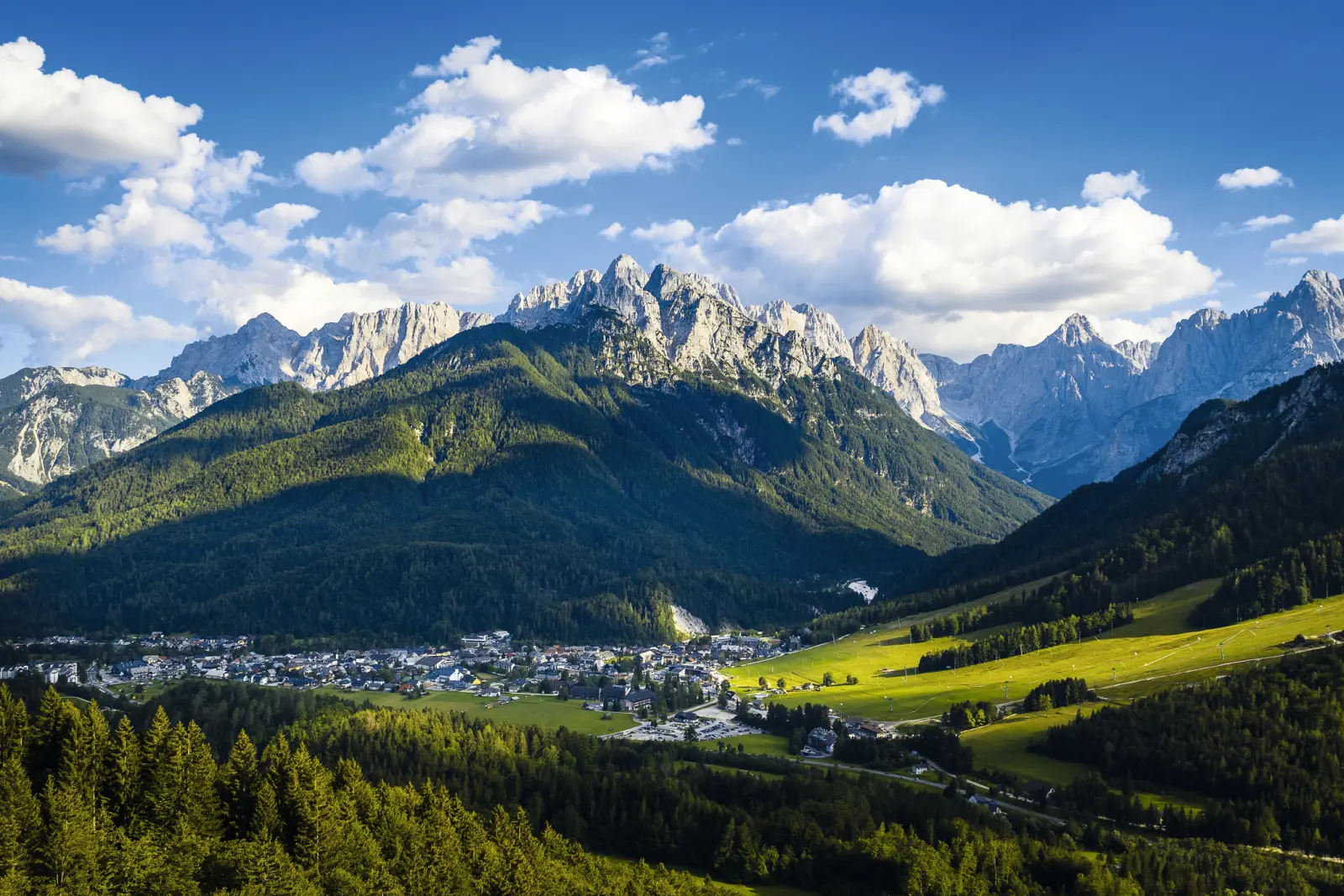 Berglandschaft mit blauem Himmel und kleinen Wolken über einem Tal mit Dorf und Wäldern.