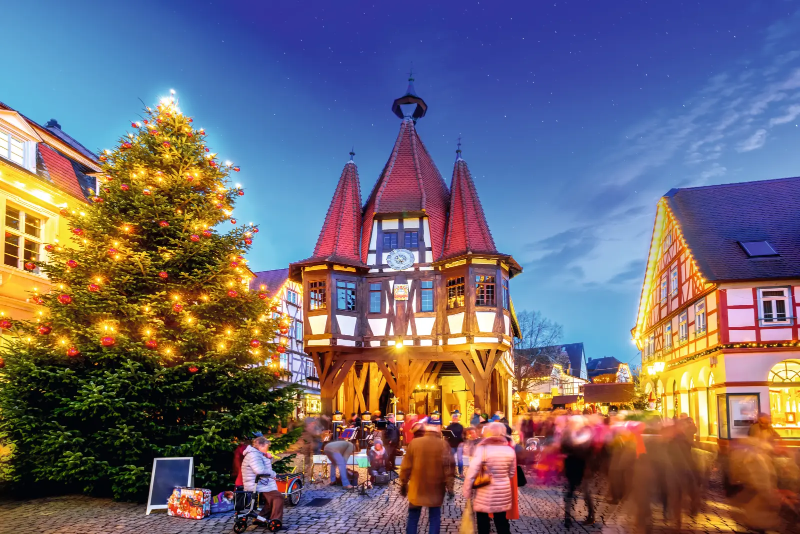 Fachwerkgebäude und Weihnachtsbaum bei Dämmerung in einem geschäftigen Platz.