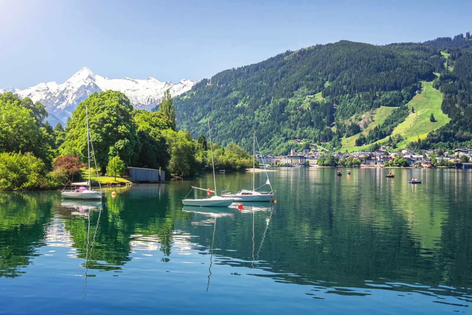 Mountain lake with sailing boats, surrounded by mountains and forest under a clear sky.