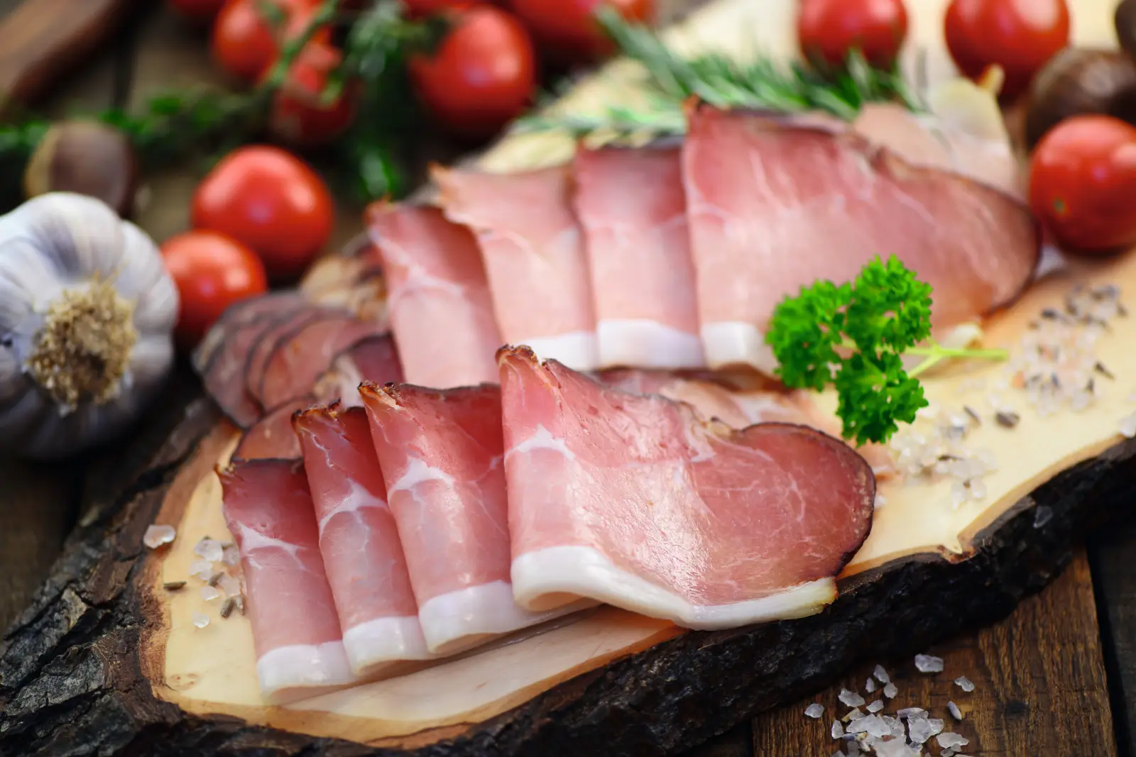 Prosciutto affettato su tagliere di legno con pomodori e aglio