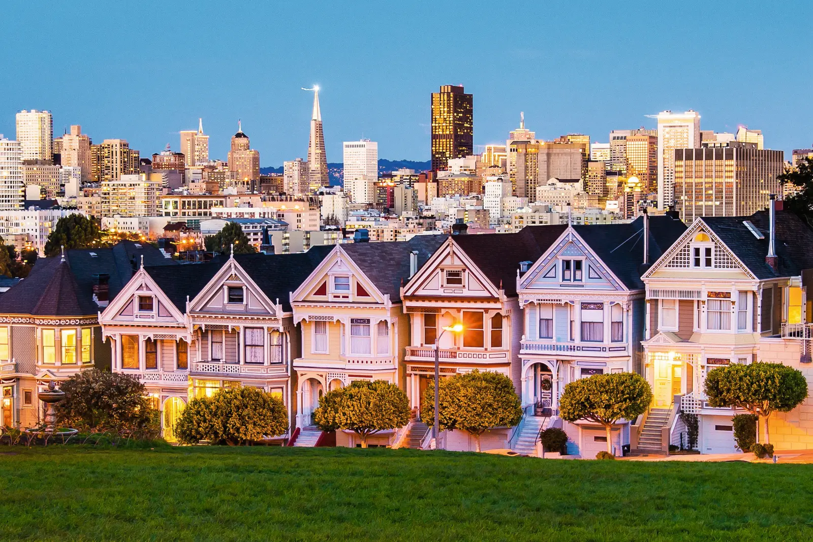 Rows of colourful Victorian houses in front of a city skyline at dusk.