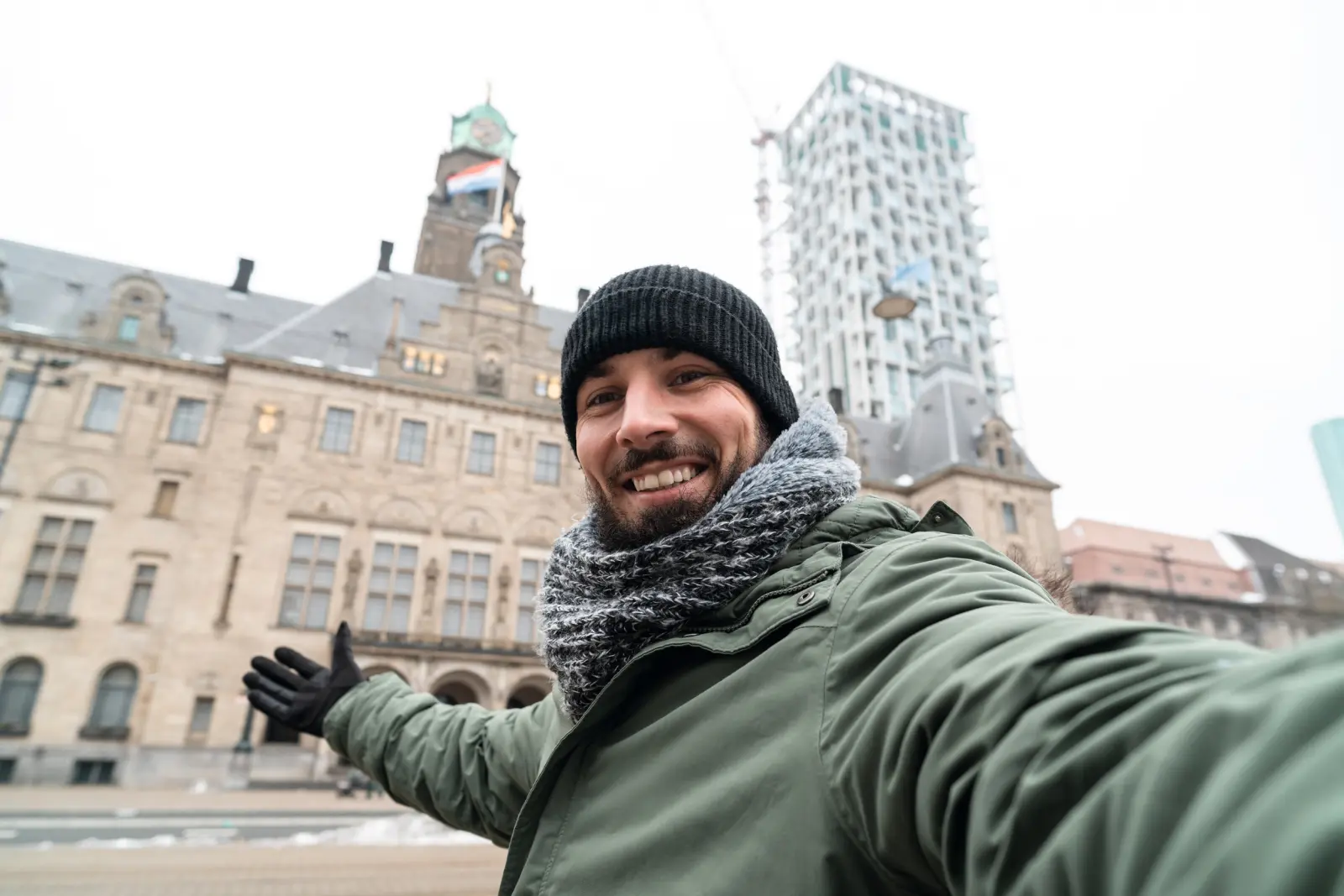 Uomo con berretto e sciarpa che si fa un selfie davanti a un edificio storico e a una torre moderna.