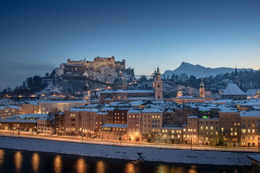 Città innevata e fortezza al crepuscolo, illuminate lungo un fiume.