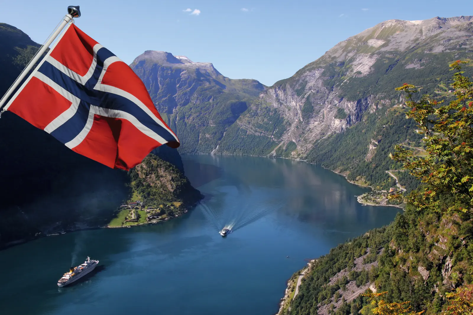Norwegian flag above a fjord landscape with boats and mountains in the background.