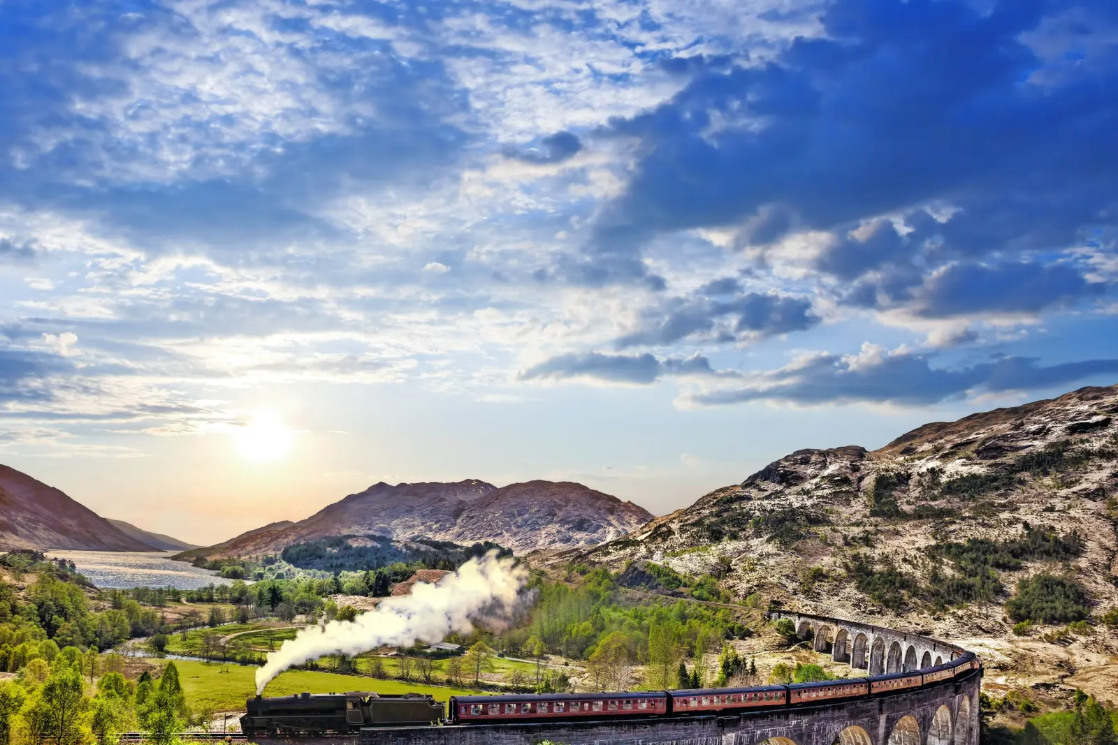 Steam engine crossing a viaduct through mountainous landscape at sunset.