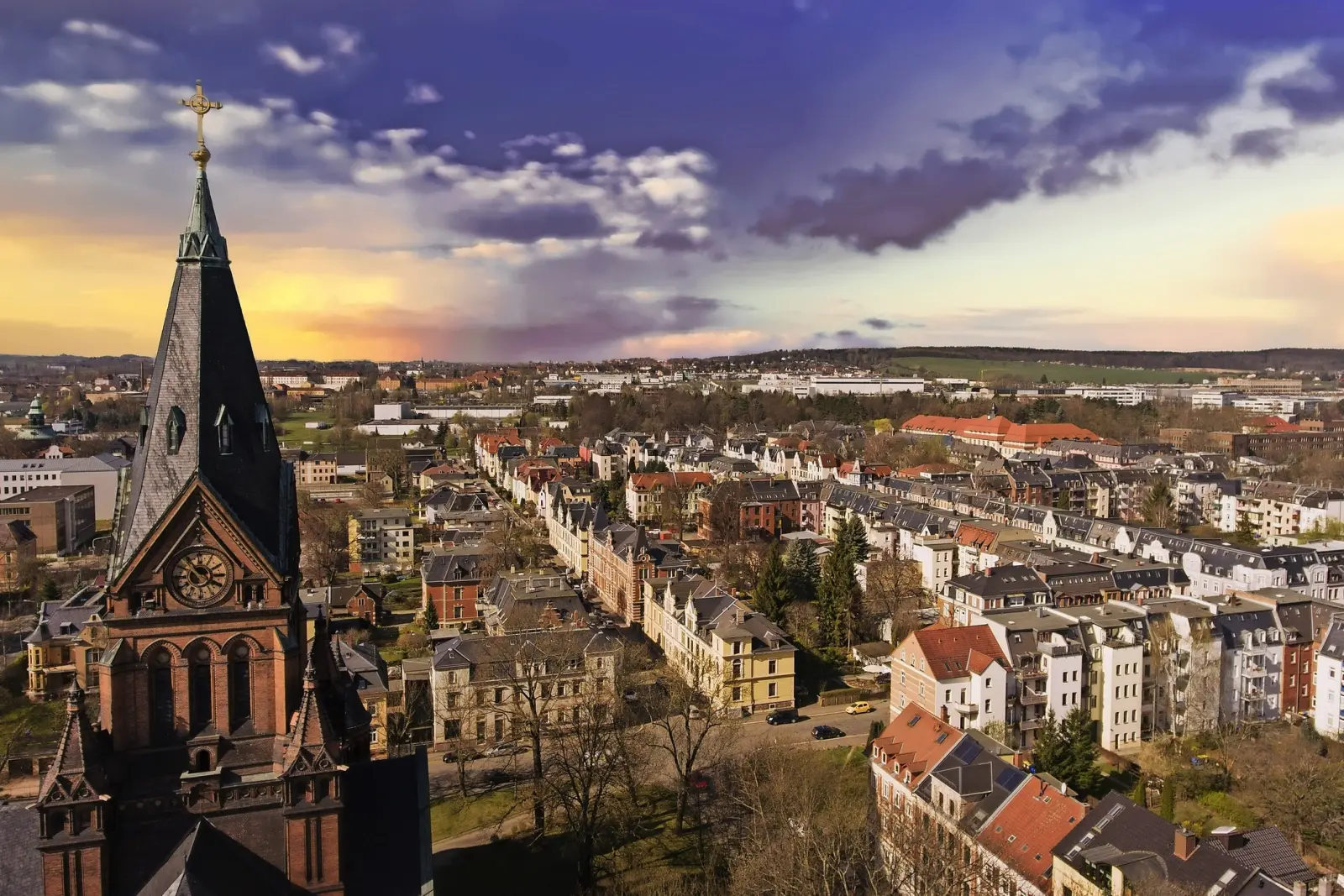Stadtansicht mit Kirchturm im Vordergrund und buntem Sonnenuntergang.