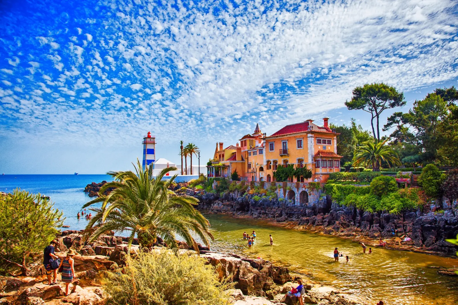 Küstenlandschaft mit Leuchtturm, Villa, Palmen und Menschen am Ufer unter blauem Himmel.