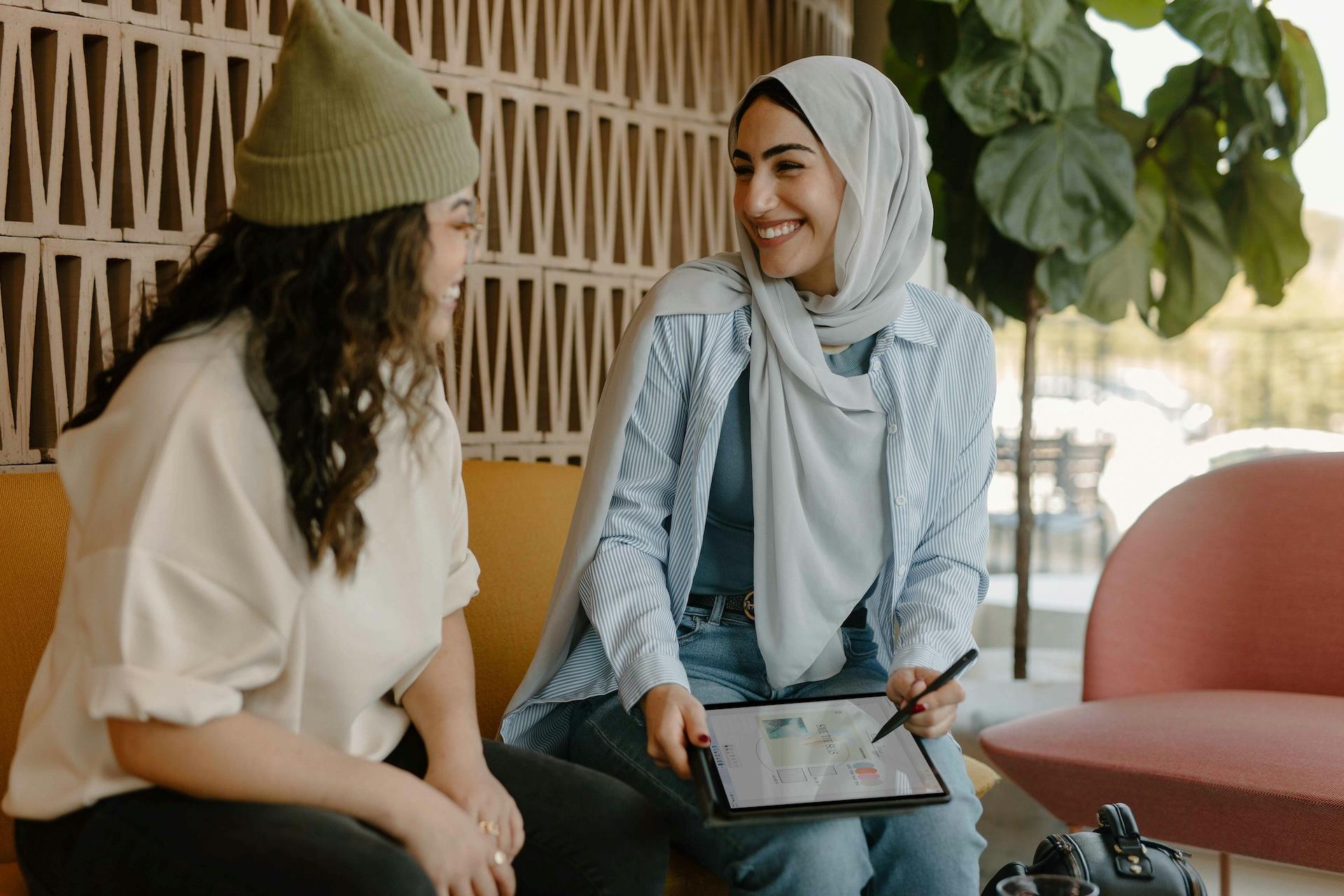 Two women engaged in a conversation, one holding a tablet and smiling, in a modern indoor setting.