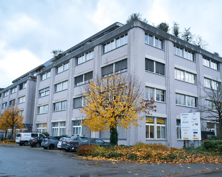 Vierstöckiges Bürogebäude mit grossen Fenstern, herbstlichen Bäumen und mehreren Autos auf dem Parkplatz davor.