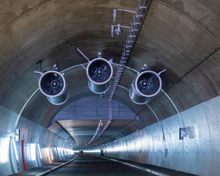 Drei grosse Lüftungsventilatoren an der Decke eines modernen Strassentunnels mit Betonwänden und Beleuchtung.