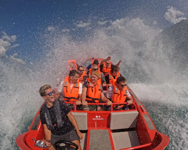 Menschen mit Schwimmwesten auf einem schnellen Jetboot, Wasser spritzt hoch, sonniger Tag, alle wirken abenteuerlustig und haben Spass.