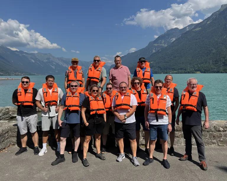 Gruppe von Menschen mit orangen Schwimmwesten steht vor einem türkisblauen See und Bergen bei sonnigem Wetter.