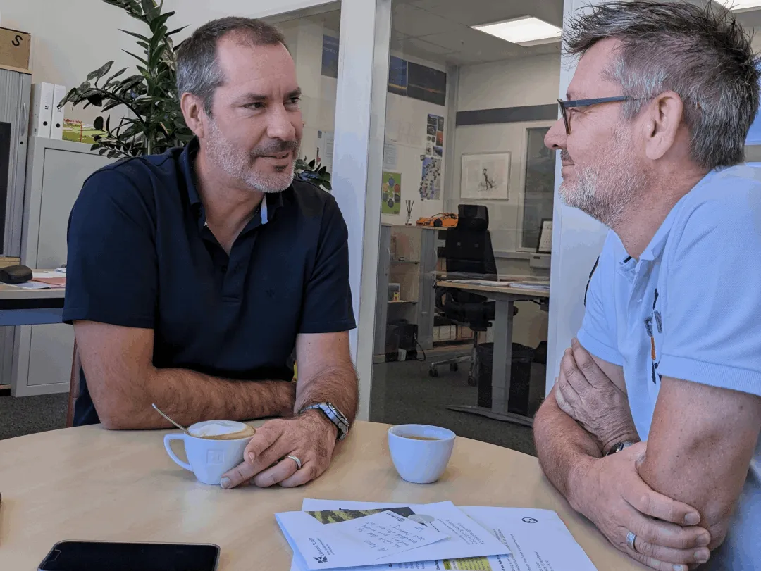 Zwei Männer sitzen an einem Tisch in einem Büro, trinken Kaffee und unterhalten sich. Auf dem Tisch liegen Papiere und ein Smartphone.