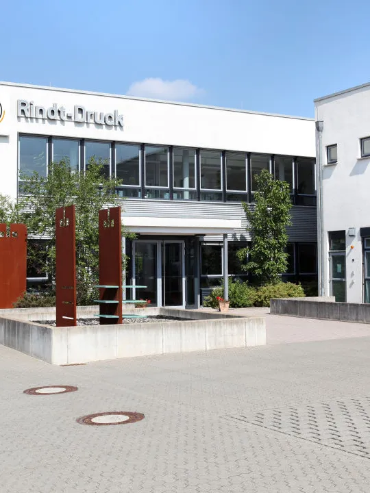 Bürokomplex der Druckerei Rindt mit Glasfassade, Eingangsbereich und Bäumen im Vordergrund, klarer Himmel im Hintergrund.