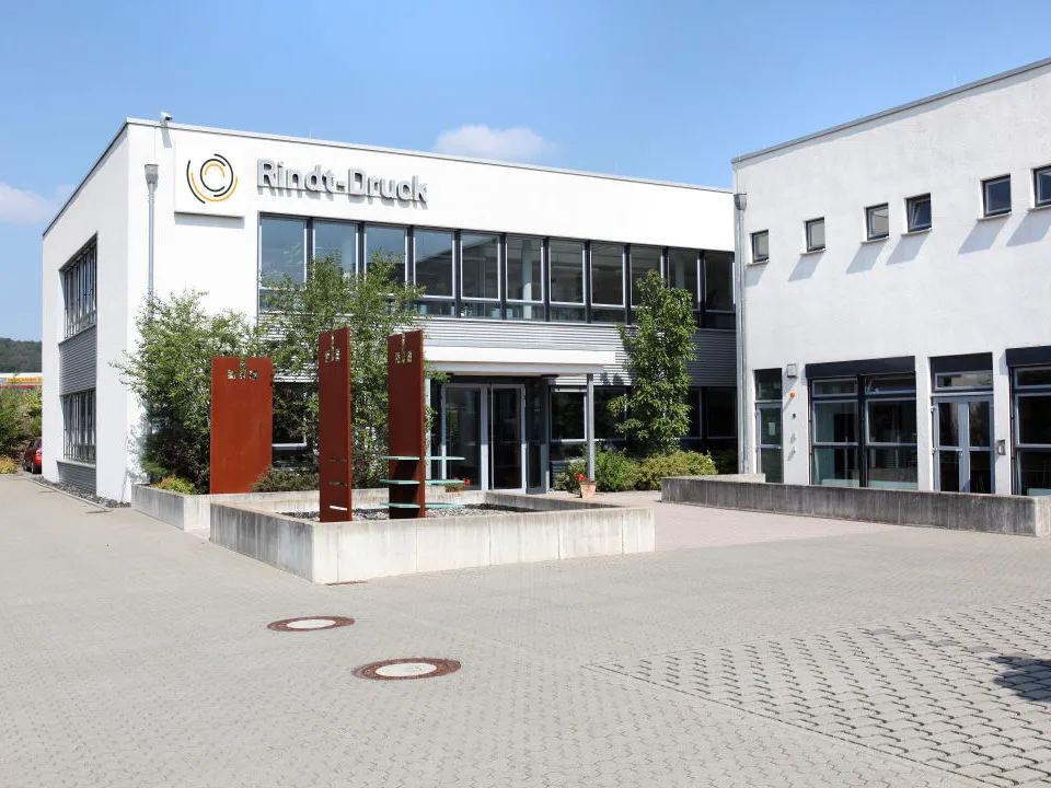 Bürokomplex der Druckerei Rindt mit Glasfassade, Eingangsbereich und Bäumen im Vordergrund, klarer Himmel im Hintergrund.